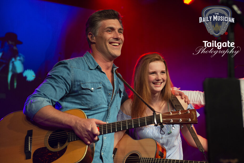 A man and a woman each hold a guitar onstage.