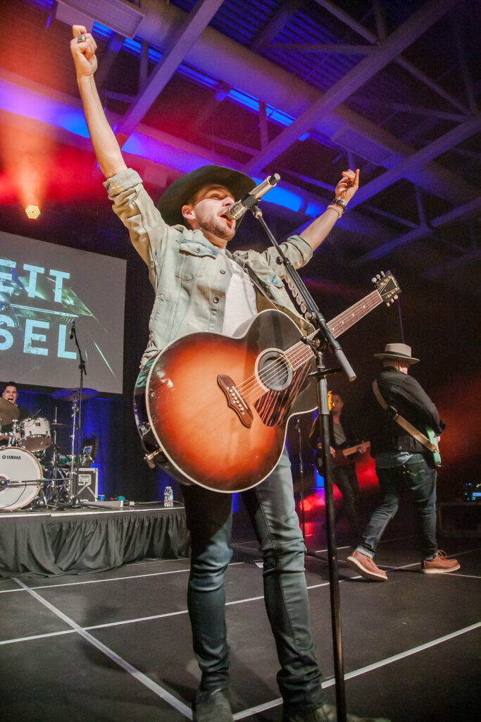 A man wearing a cowboy hat and a guitar raises his arms in the arm and sings into a microphone.