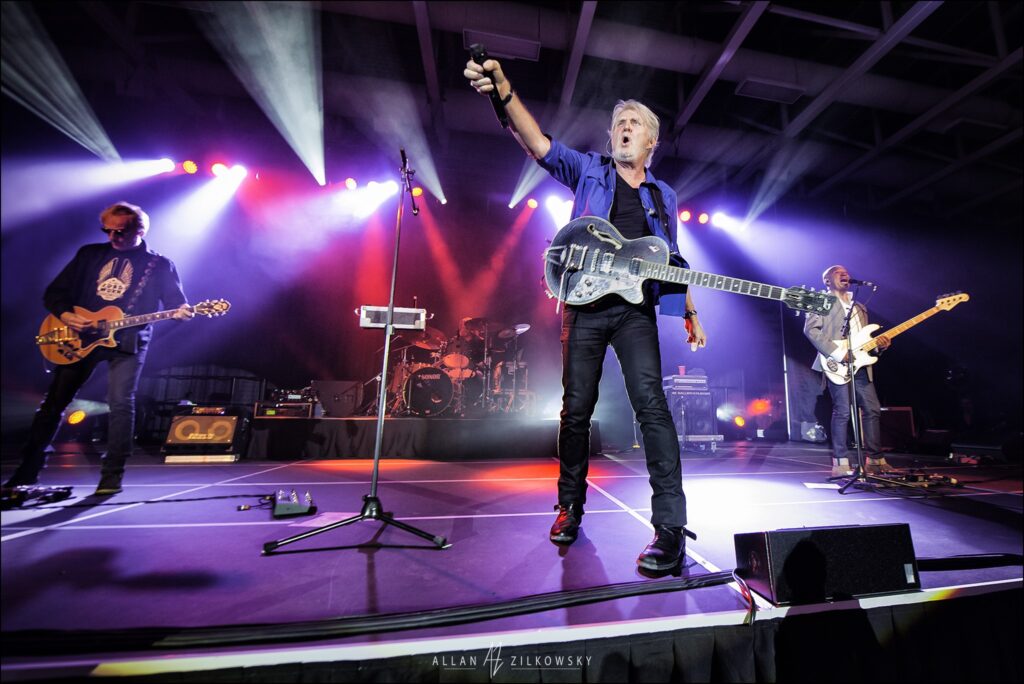 Tom Cochrane plays guitar on a stage.