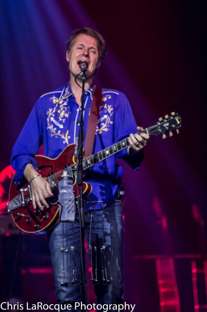 A man in a Western-style shirt plays guitar and sings on stage.
