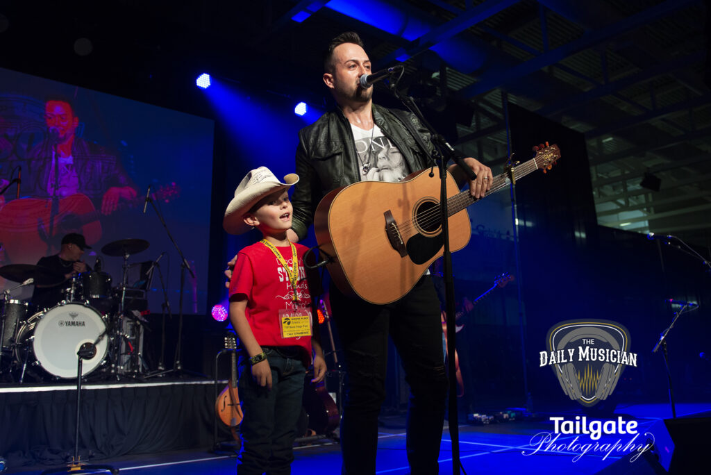 A man with a guitar stands on the stage, with a young boy in