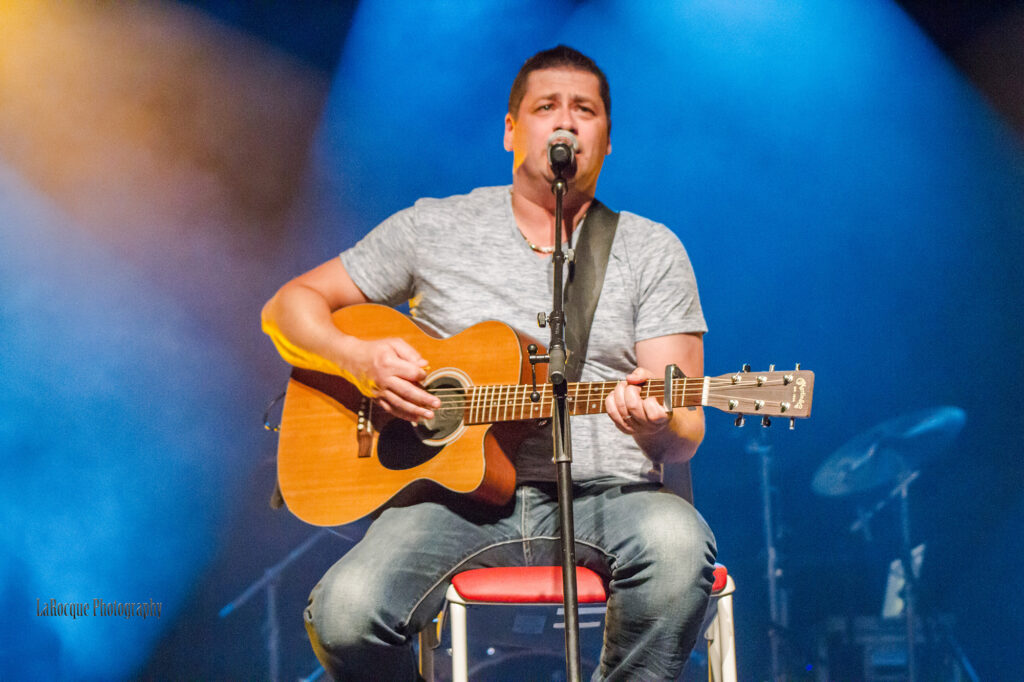 A man in a plain t-shirt sits on a stool and plays the guitar, while singing into a microphone on stage.