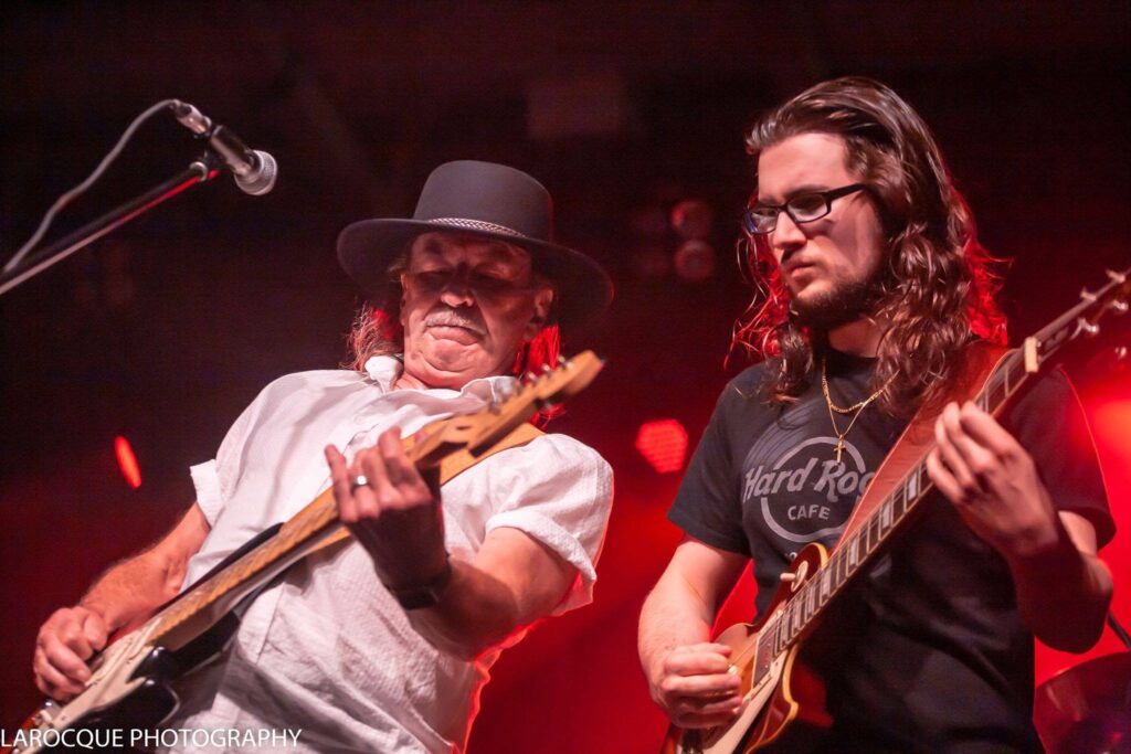 Two men play guitars on stage. One wears a black hat, the other has long hair and glasses.
